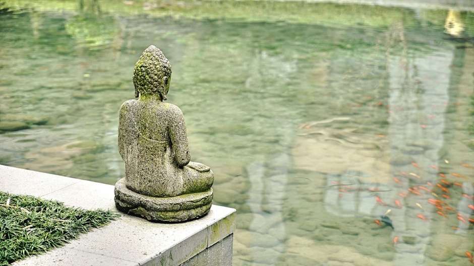 Looking at the back of a Buddha Statue overlooking Koi swimming in a pond